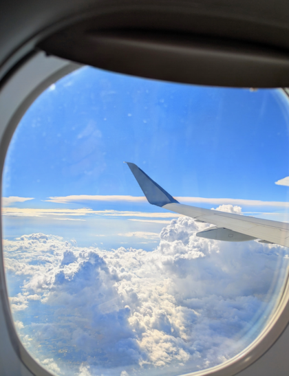 a blue sky above an airplane wing. Below, ornate clouds. Far below, farmland.